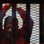 Deposed President Mohamed Mursi greets his lawyers and people from behind bars at a court wearing the red uniform of a prisoner sentenced to death, during his court appearance with Muslim Brotherhood members on the outskirts of Cairo, Egypt