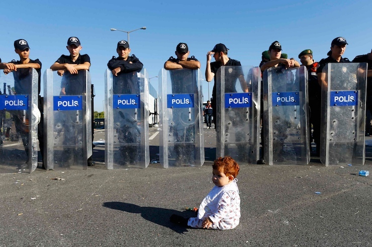 An infant sits on the road in front of a line of Turkish police who block migrants on a highway near Edirne