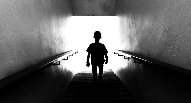 Silhouette of a person walking up a dim stairwell toward bright light at the top.