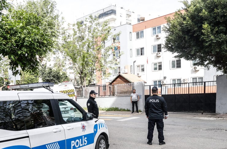 Police car with blue stripe labeled POLIS on a residential street as two officers stand near a gated apartment building and a man watches from the sidewalk.