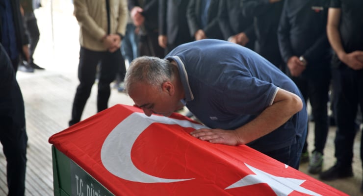 Man kissing a coffin draped in a Turkish flag at a funeral, surrounded by mourners and onlookers.