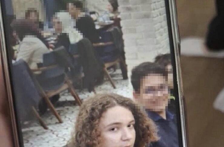 Portrait of a young person with curly hair looking at the camera in a cafe, with people seated in the background.