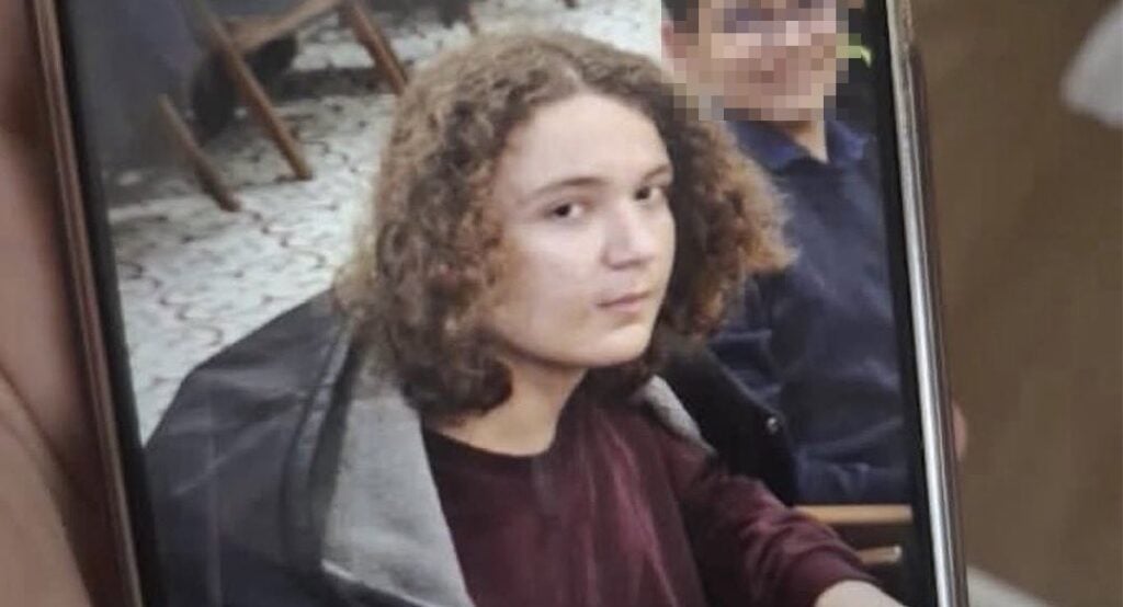 Portrait of a person with curly hair looking at the camera while seated indoors, with another person blurred in the background ( photo on a phone screen ).