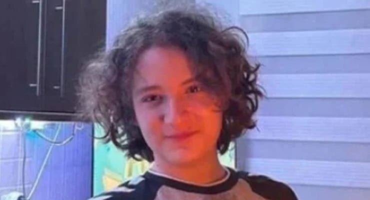 Child with curly hair facing the camera, wearing a gray and black shirt, indoors near a blue wall and a fish tank in the background.