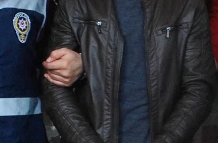 Person in a brown leather jacket with hands cuffed in front, escorted by a police officer in a blue uniform.