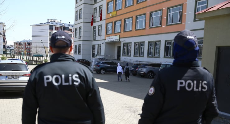 Two police officers wearing POLIS jackets stand in a paved courtyard in front of a multi‑story building with flags while pedestrians walk nearby.