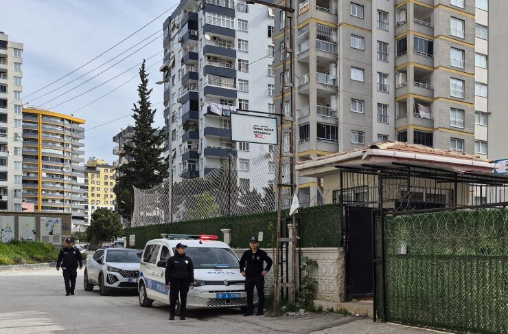 Police officers stand near a blue-and-white patrol car with flashing lights in a residential area with tall apartment buildings and a gated complex behind them.