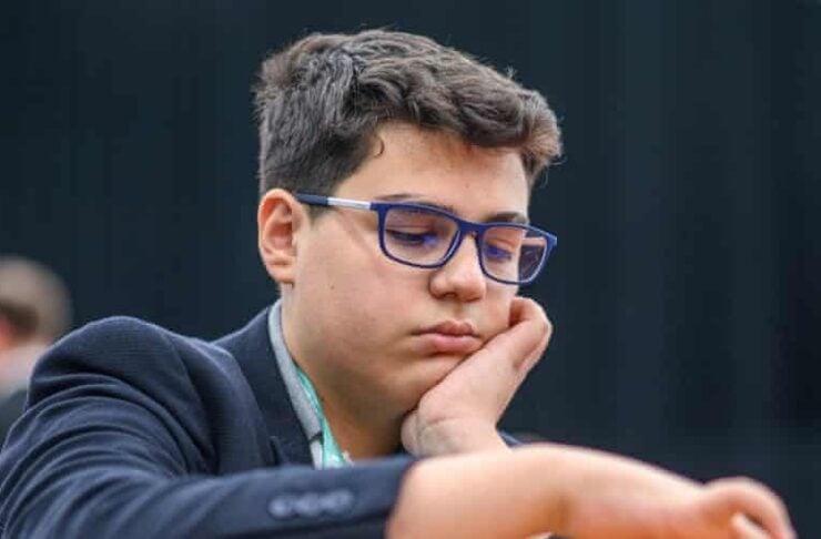 Boy with blue glasses concentrating on a chessboard, resting his chin on his hand as he contemplates a move.