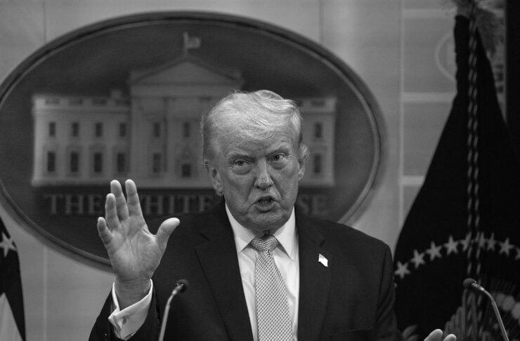 Black-and-white photo of a suited man at a presidential podium, with one hand raised as he speaks, White House seal in front.