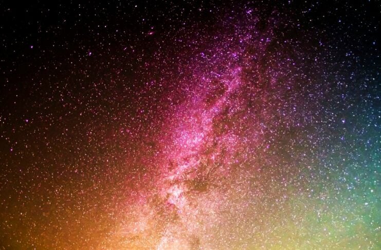 Silhouette of a person standing on a rocky hill under a colorful Milky Way with countless stars.
