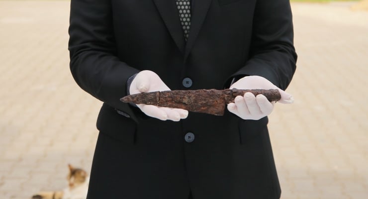 Person in a black suit and white gloves presenting a long, rusty iron artifact outdoors.