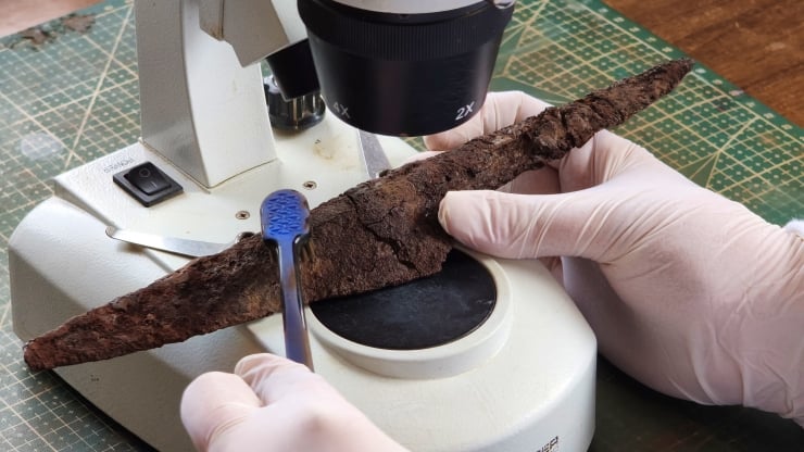 Gloved hands holding a long, cracked fossil or rock specimen under a microscope with a cutting instrument nearby.