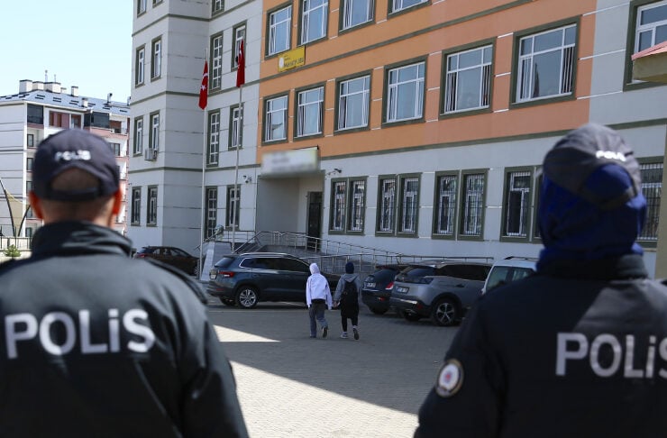 Two police officers face away from the camera, watching a group of pedestrians approach a multi-story building with Turkish flags, parked cars along the side.