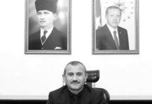 Man in a suit seated at a desk in an office, hands folded, with a small Turkish flag on the desk and two framed portraits on the wall behind.