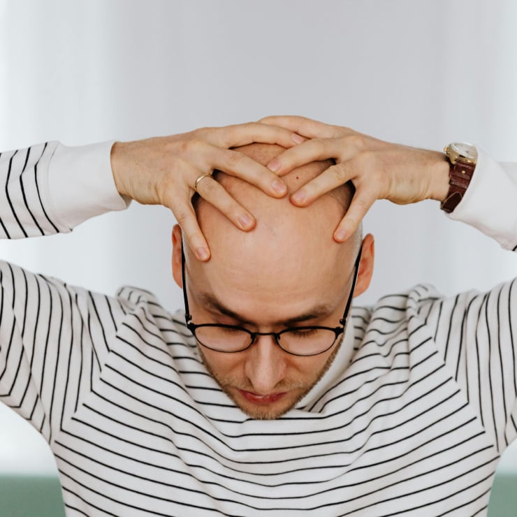 Man wearing glasses holding his head with both hands, looking down in apparent stress or headache relief posture.