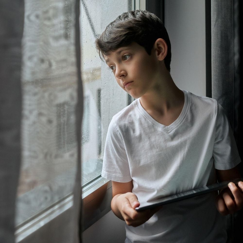 Boy standing by a rainy window, looking out pensively while holding a tablet.
