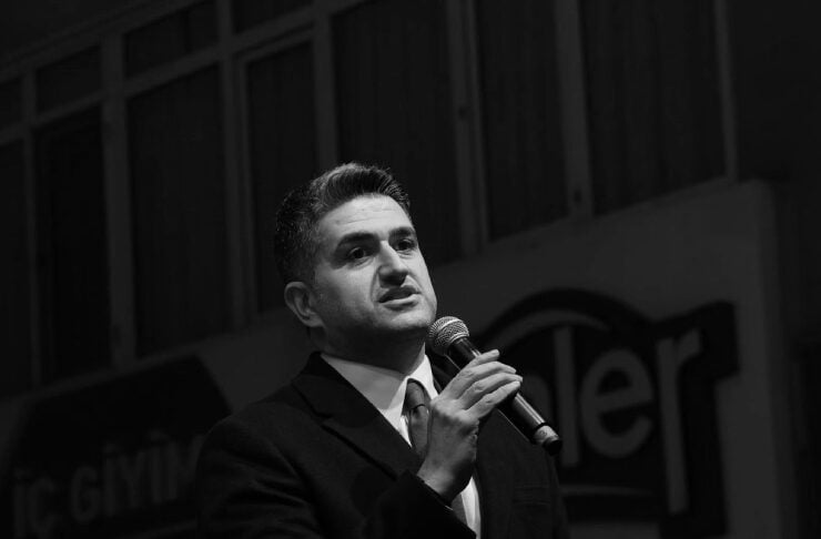 Man in a dark suit speaks into a handheld microphone on a stage.