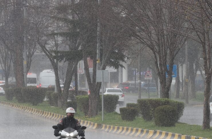 Motorcyclist wearing a white helmet rides a scooter through heavy rain on a city street with wet pavement and cars in the background