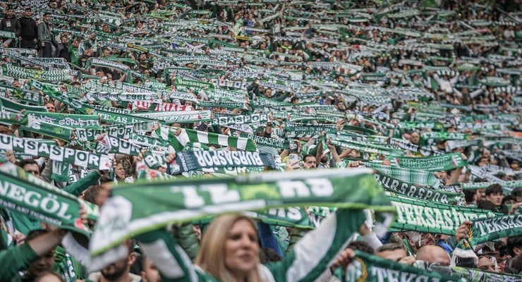 Large crowd of football fans in green and white scarves waving at a stadium, celebrating.