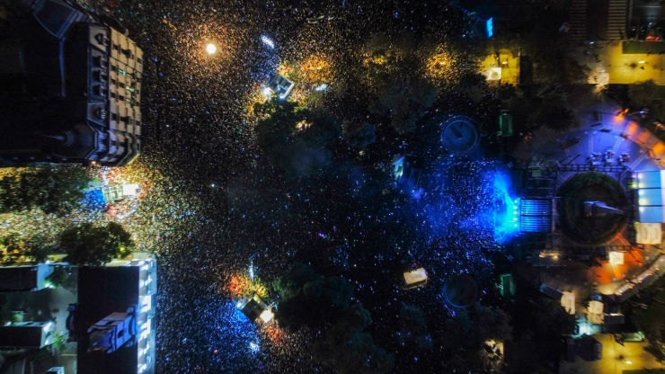 Aerial night view of a crowded city square with colorful stage lights, confetti, and surrounding lit buildings and trees.