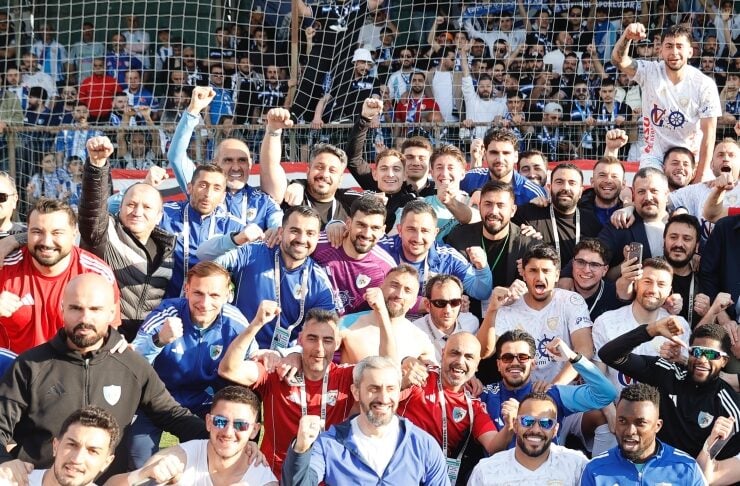 Large group of male soccer players and fans celebrating on a field with a net behind them, many fists raised in victory.