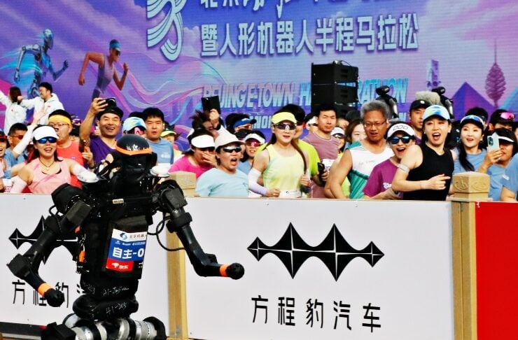 Humanoid robot performing a dance on a race track as cheering spectators watch from behind a barrier with banners in the background.