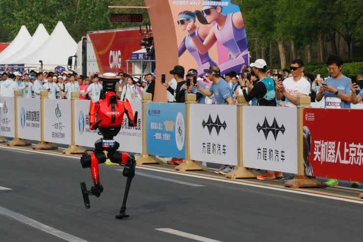 Red humanoid robot running on a city street during a marathon event, with spectators behind barriers and race banners in the background.
