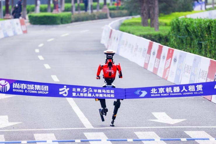 Red bipedal robot breaks through a blue finish-line banner on a city street race course, arms raised as it runs toward the camera.