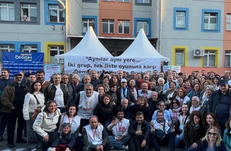 Large group of people posing for a group photo on a city street in front of a white tent and banner that reads: 'Aynılar aynı yere... iki grup, tek liste oyununa karşı'.