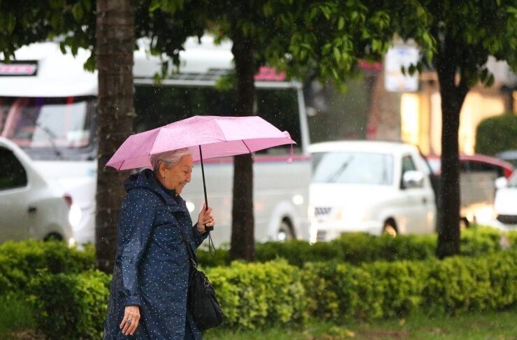 Older woman wearing a navy raincoat with a pink umbrella walking on a wet street in the rain.