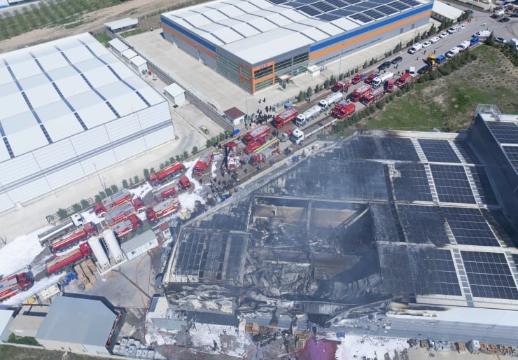 Aerial view of a burned warehouse with smoke, surrounded by fire trucks and emergency crews.