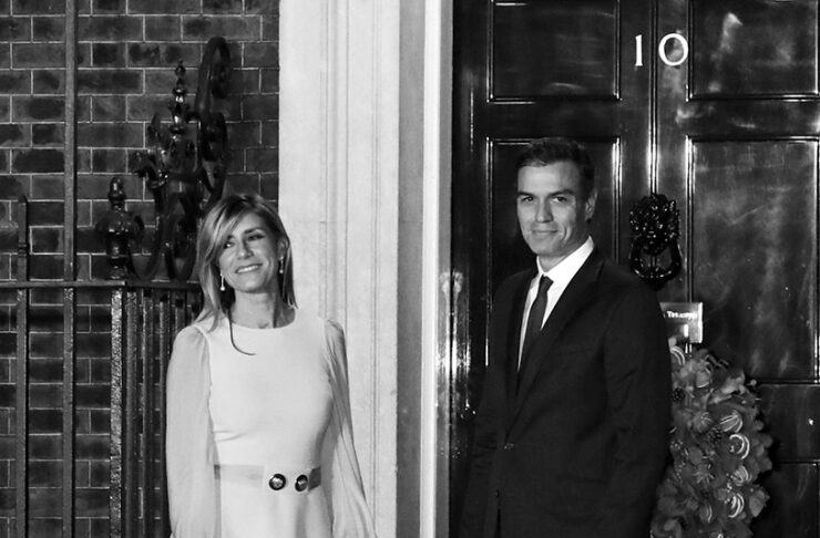 Man in a dark suit and woman in a light dress stand on a doorstep in front of a black door marked 10, smiling.