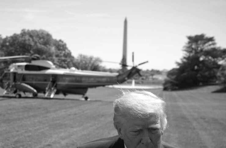 Man in a dark suit and tie stands on a grassy airfield with a helicopter in the background and a tall obelisk-like structure nearby, looking downward with a serious expression, wearing a small flag pin on the lapel.
