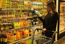 Woman in a black coat pushes a shopping cart while picking up a bottle from the refrigerated juice/snacks aisle in a supermarket.