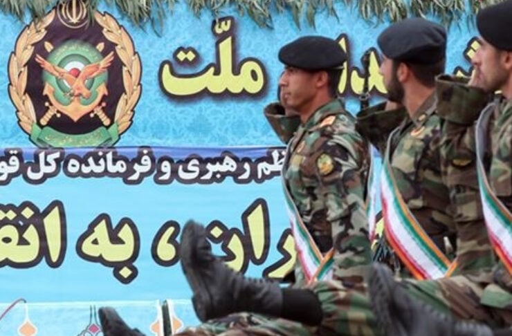 Soldiers in camouflage uniforms marching in formation with raised legs in a parade, against a blue banner with an emblem and Persian text in the background.