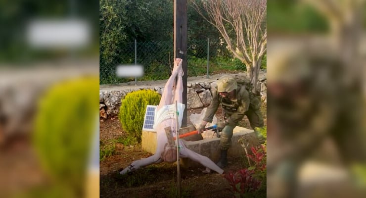 Life-size mannequin suspended from a vertical pole outdoors, with a solar panel on its chest, while a person in uniform works with a tool nearby.