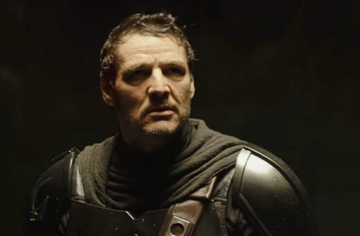 Man wearing dark armored suit with shoulder plates and a brown strap, standing in dim light.