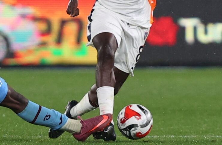 Two players compete for a soccer ball on a green field, one wearing white shorts and pink cleats, the other in blue socks, during a tackle.
