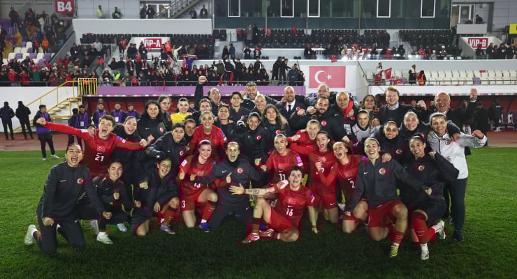 Turkish women’s soccer team posing for a group photo on the field after a match, in red uniforms, with fans and banners visible in the stands behind them.