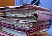 Person holding a tall stack of tangled manila folders and papers in both hands.