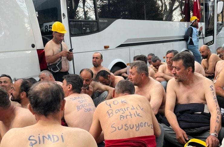 Group of shirtless men sitting on the ground beside a large bus, several with handwritten messages on their backs in Turkish.