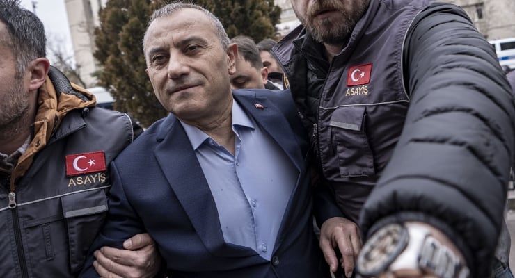 Man in a blue suit is escorted by two security officers wearing jackets with Turkish flags and 'ASAYIŞ' patches.