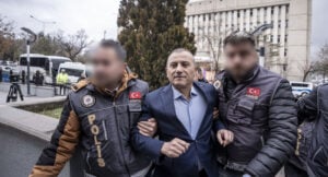 Man in a blue suit being escorted by two police officers wearing 'POLIS' jackets with Turkish flags in an urban area. Central figure looks forward as officers hold his arms.