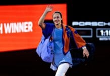 Female athlete in blue outfit waves while carrying sports bags, walking off a dark court with a red
