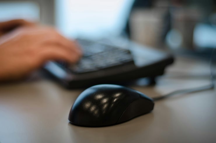 Close-up of a black computer mouse on a desk, with a person typing on a keyboard in the blurred background