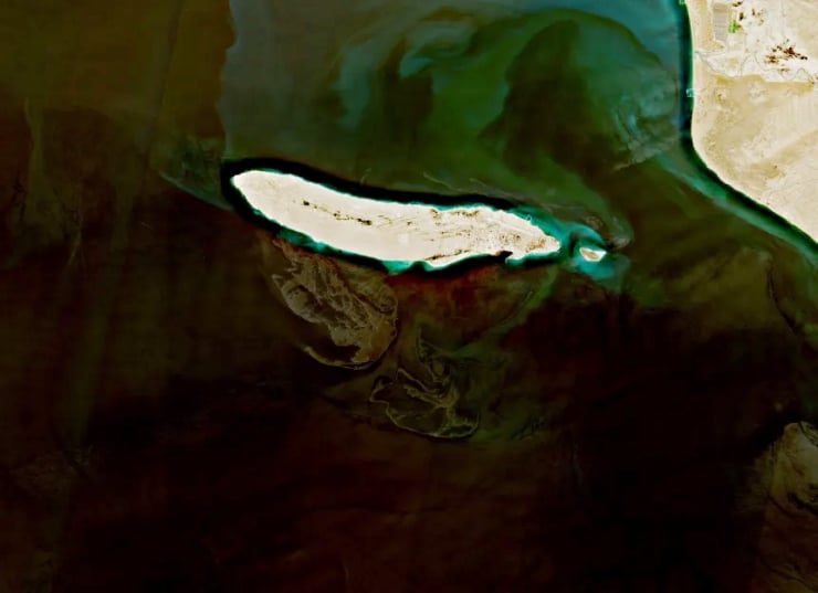 Aerial view of a long white sandbar or island jutting into pale turquoise water with dark surrounding sea and rocky formations nearby.