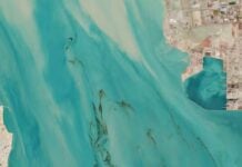 Aerial view of turquoise coastal water with light currents, showing land on the top and left edges and dark water channels across the sea.