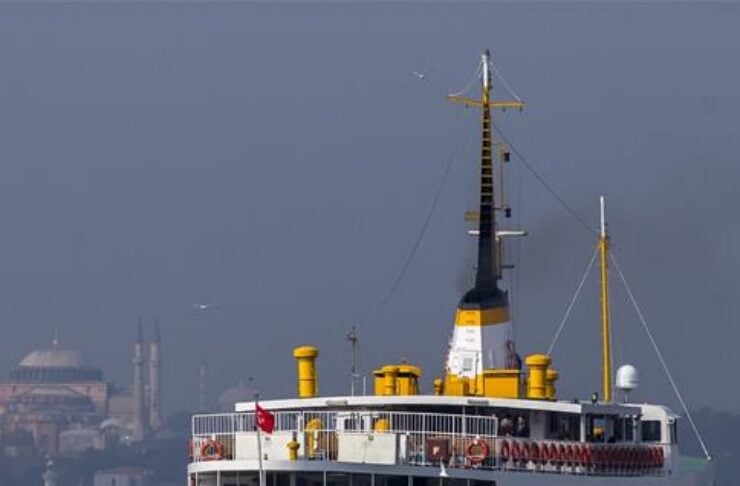 Passenger ferry with white and yellow hull sailing in a harbor, city skyline in the background.