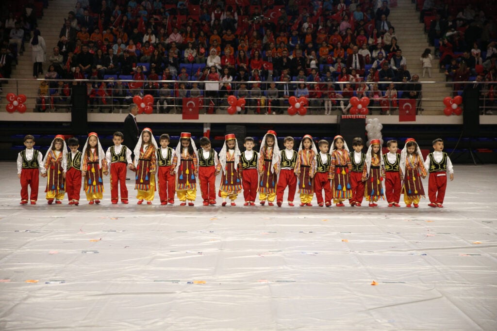 Group of children in colorful Turkish folk costumes standing in a line on a performance floor, with a large audience and Turkish flags in the stands behind them.