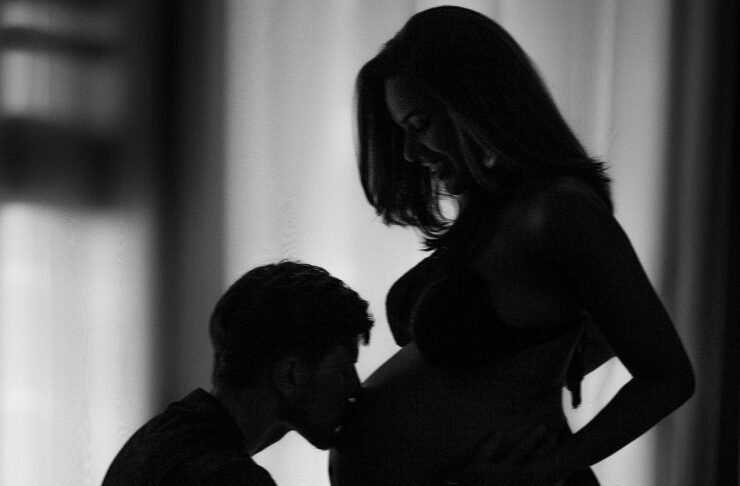 Silhouette of a pregnant woman standing as her partner kneels to kiss her belly.